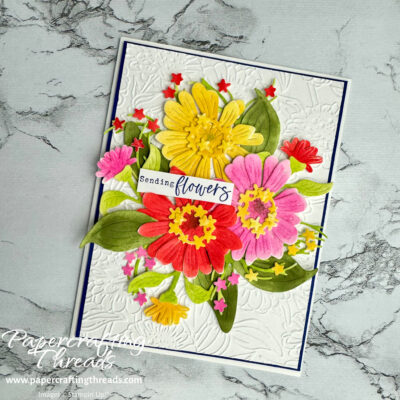 Colorful yellow, red, and hot pink zinnia flowers and buds with green foliage rest on an embossed white card front with hint of dark blue border