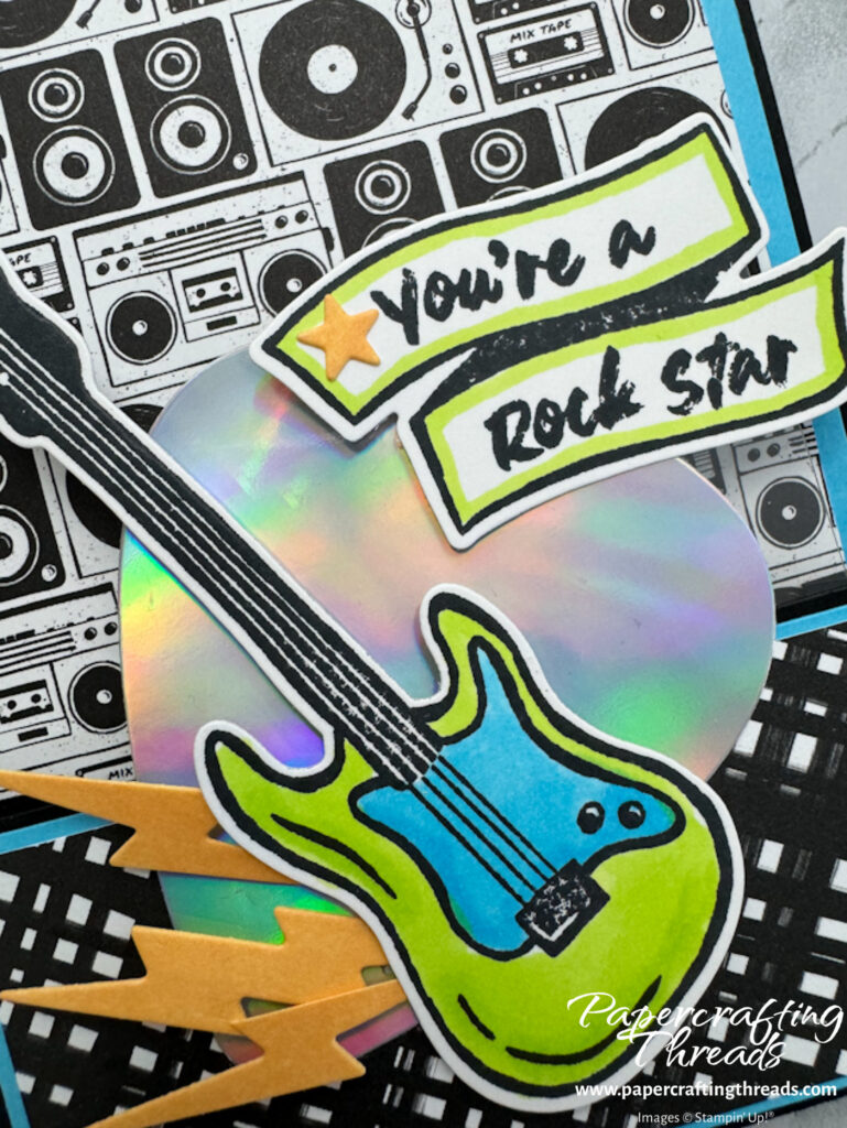 Close up of turquoise and lime green electric guitar over a holographic extra large guitar pick with orange lightning bolts shooting from the side and sentiment of you're a rock star on a banner.