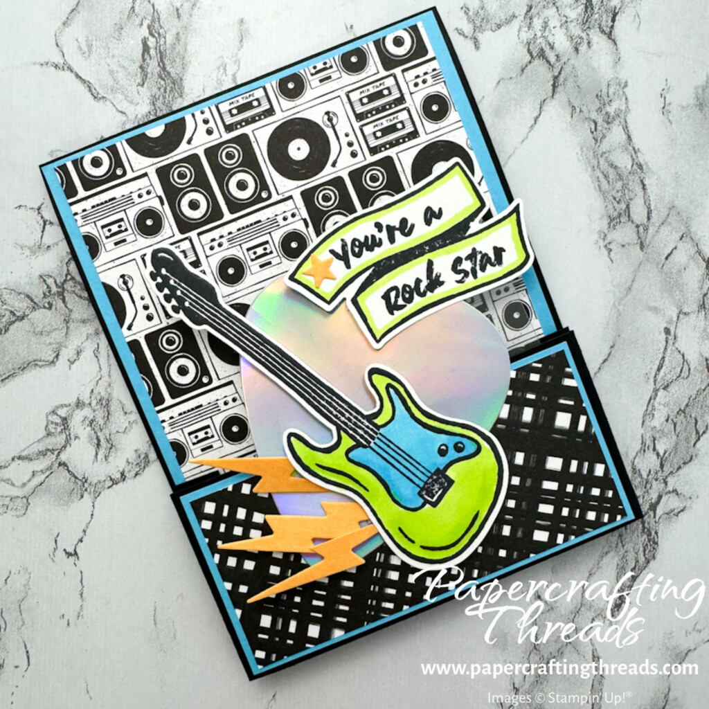 Front view of Single Bridge Card with patterned black and white background over turquoise cardstock with bright turquoise and lime green guitar, orange lightning bolts, holographic giant sized guitar pick and banner sentiment of 'you're a rock star'