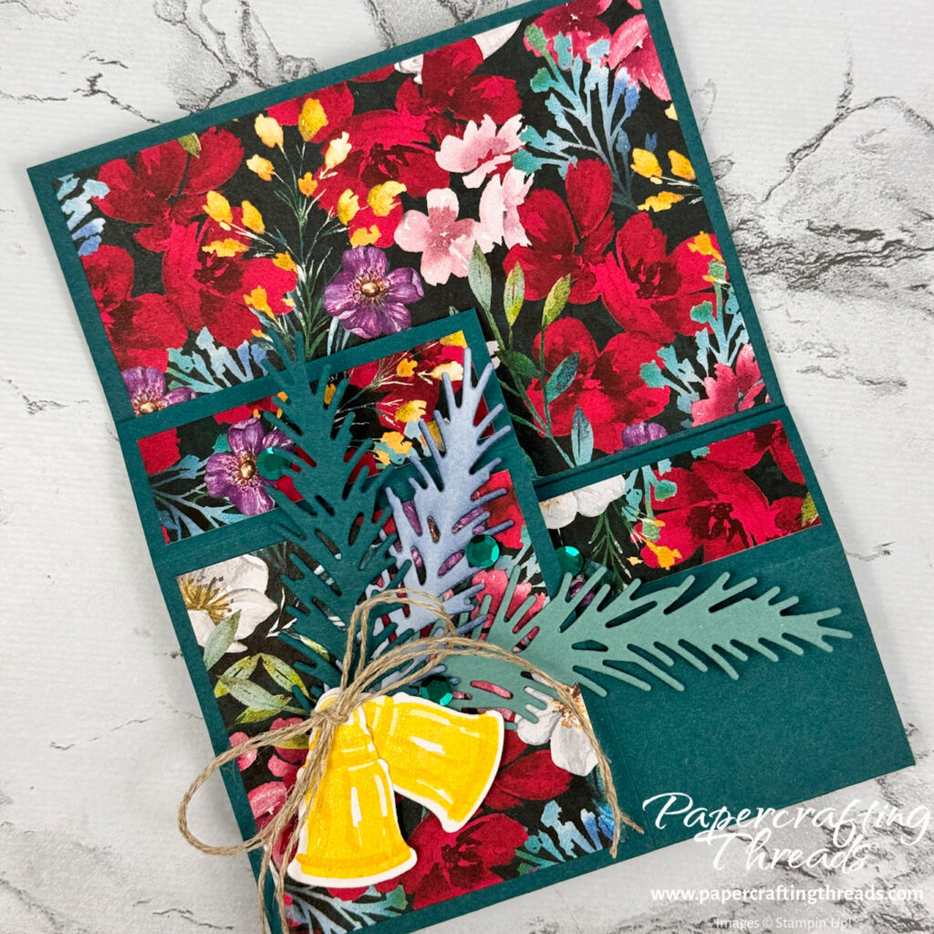 Folded Flat card with brilliant floral pattern in reds, purples, pinks, gold, and blues on a Pretty Peacock base with gold bells, pine boughs and a linen thread bow