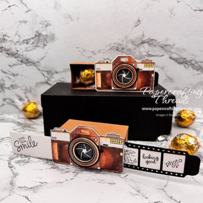 Two Matchbox Double Slider Cameras shown. Brown stamped and die cut camera images adhered together to create a realistic 3D camera attached to a brown matchbox drawer that holds chocolate truffles. One camera shows the secret messages revealed on each side. Greeting of 'love this smile' extends on white strip to the left. Film strip extends to the right with three cells, 'photo op', 'looking good' and a beach scene. Second camera is shown with the hidden drawer open and filled with truffles.