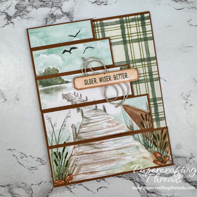 Staggered Z-Fold or Staircase Card. Front features a scenic pier extending into a lake with shoreline cattails, and a trio of birds flying high in the sky. The scene is cut into strips and staggered along the side, creating a staircase effect. A 'Older, Wiser, Better' greeting spans the middle with a nest of linen thread behind it.