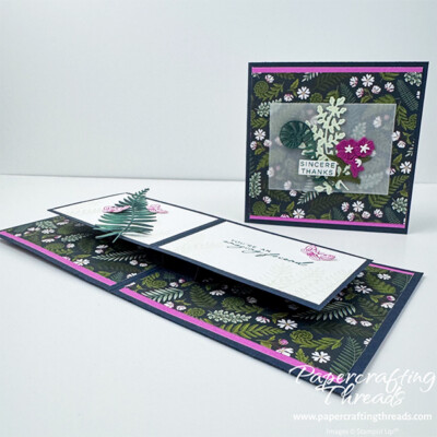Front view of Butterfly Effect Fun Fold Card. Secret Sea colored square card base, with top and bottom strips of Petunia Pop, a fern and flora patterned paper in deep blues, greens and touches of white and magenta flowers. Focal point set on a piece of vellum consists of die cut fiddle head fern, foliage and a hot pink floral element. Sentiment is sincere thanks. Inside of card opens to reveal a floating pop up similar to butterfly wings. Decorated with more ferns and pink moths.