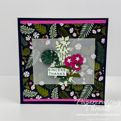 Front view of Butterfly Effect Fun Fold Card. Secret Sea colored square card base, with top and bottom strips of Petunia Pop, a fern and flora patterned paper in deep blues, greens and touches of white and magenta flowers. Focal point set on a piece of vellum consists of die cut fiddle head fern, foliage and a hot pink floral element. Sentiment is sincere thanks.