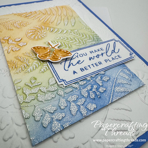 Embossed Spotlight Technique Card features a card base in Hydrangea Hue, a pretty bluish color with just a hint of purple. Ferns and flora are dry embossed into a layer of white cardstock. A large rectangular focal point is dry embossed and colored in swaths of Golden Glow, Soft Sea Foam and Hydrangea Hue colors, with the edges blending into each other. A Golden Glow moth alights on the greeting 'you make the world a better place'.