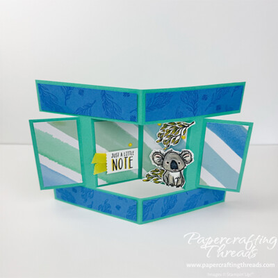 Pop Out Burst Fun Fold Card Front. A bright turquoise card base with a full middle panel that pops out of the center of the card. A koala nestled in eucalyptus branches sits on the right panel. Greeting of 'Just a Note' with a yellow ribbon behind it is on the opposite panel.