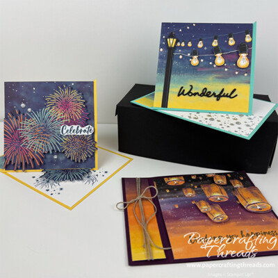 Twisted Easel Trio Alternatives May 2026 Paper Pumpkin Kit Sparkling Celebrations. Three cards shown. Top right is on a turquoise base with purples, blues and yellow evening sky. Strings of lightbulbs lead to a lamppost. Large sentiment of 'wonderful' at the bottom. Card on lower right shows a purple and orange evening sky with floating lanterns rising up. Sentiment is Wishing you happiness. Card on far left has a yellow base and an evening starry sky with fireworks in oranges, yellows, reds and turquoise. Sentiment is Celebrate.