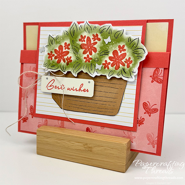 Easy Little Fun Fold card alternative for Bikes & Blooms Paper Pumpkin kit. Card is shown closed. Front has white flap extending down from the top of the landscape card. A tan woven basket full of flowers fills the flap. Sentiment is best wishes with a linen thread bow. A coral colored lower flap that flips up under the upper flap is covered in tone on tone little butterflies.