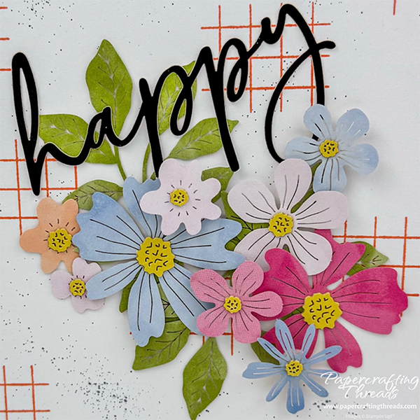 Closeup view of a white card base with watercolored floral die cuts in peach, soft pink, soft blues and soft purples. A large black greeting of 'happy' is nestled above a large spray of florals and greenery with a cross hatch of orange in the background and black splatters of ink.