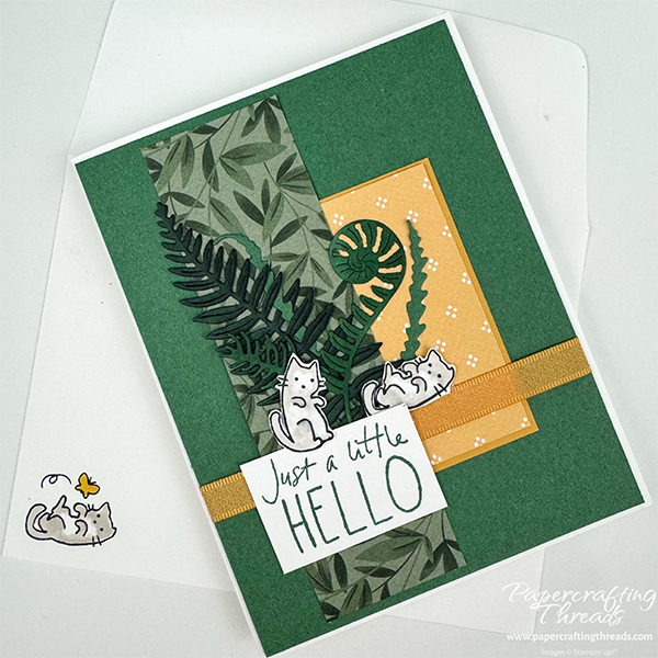 Step Up Your Cardmaking sample card with two patterned papers, one leafy greens and one neutral golden pattern layered over pine green cardstock and white card base. Multiple die cut fernlike foliage in various shades of green gathered together at base. Gold ribbon is wrapped around bottom third. Two playful kittens nestled around large sentiment of 'just a little HELLO'. Matching envelope with kitten playing with butterfly in lower left corner.
