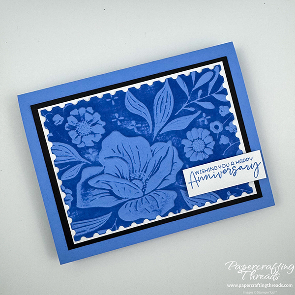 Card showcasing an embossing folder technique has large floral image debossed with blue ink on a raised background. White mat on top of black mat and all mounted on blue card base. Sentiment is 'wishing you a happy anniversary'.