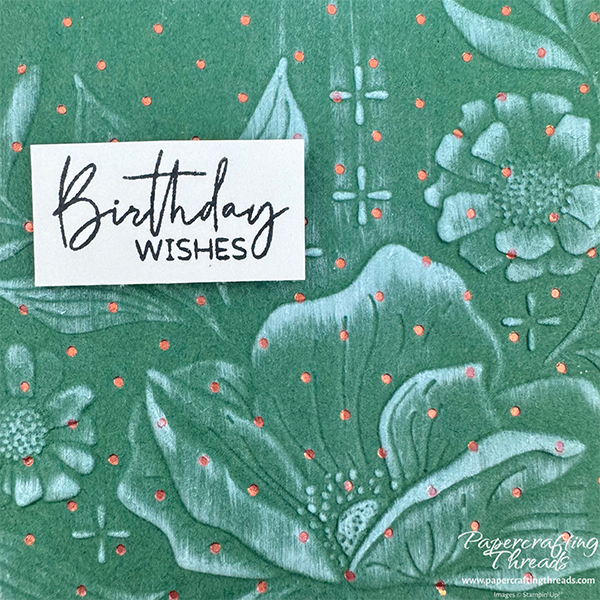 Close up of foiled copper dots over a green background with floral images raised embossed, and inked with white pigment ink. Sentiment is 'Birthday wishes'.