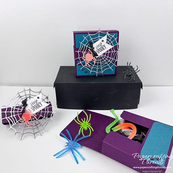 Multiple views of Web of Fun Treat Box with Hot Foil die cut. Deep purple and turquoise treat box with silver foiled die cut web. Bright orange shellacked spider with greeting tag of 'very spooky' accented with tiny black ribbon knot and purple dots. Shown closed and opened with halloween treats spilling out and belly band off to the side.