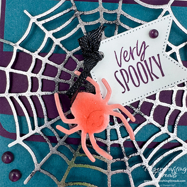 Close up of Web of Fun Treat Box with Hot Foil die cut. Deep purple and turquoise treat box with silver foiled die cut web. Bright orange shellacked spider with greeting tag of 'very spooky' accented with tiny black ribbon knot and purple dots.